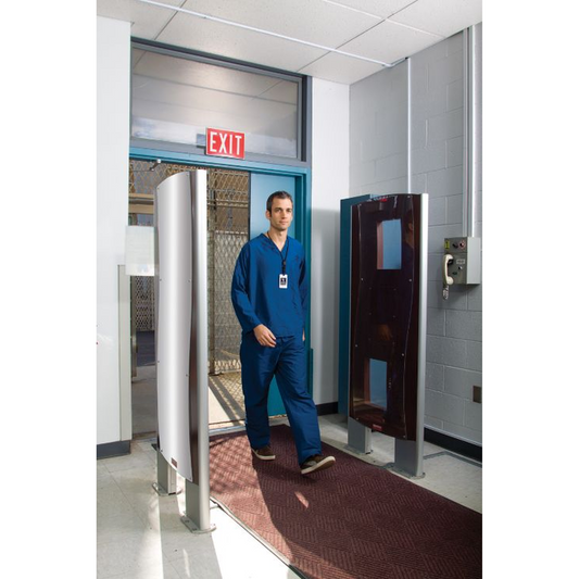 Person in blue scrubs standing in a medical setting with a metal detector and exit sign.
